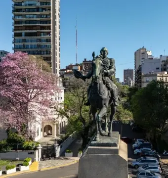 Bartolomé Mitre Monument with a dark equestrian statue on a grey pedestal, framed by city buildings and a blooming tree.
