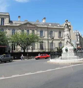 The Gavarni Fountain stands on a street corner behind a black metal fence, with sculpted figures, parked cars, and nearby buildings under a blue sky.