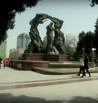 Huseyn Javid Monument with a central standing figure and two arcing bronze figures over a red base beside a fountain in a park.