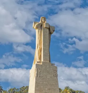Monument to Columbus, a tall stone sculpture on a rectangular base in an open plaza with trees and a blue sky.