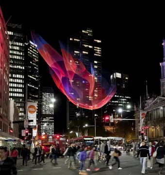 A large pink, red, and blue mesh sculpture hangs above a busy Sydney intersection at night with pedestrians and city traffic below.