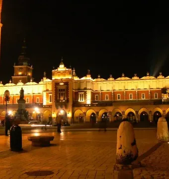 Adam Mickiewicz Monument lit at night in a city square with an arcaded building and clock tower behind it.