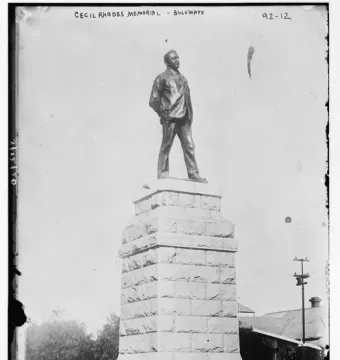 Statue of Cecil Rhodes standing atop a tall stone plinth outdoors.