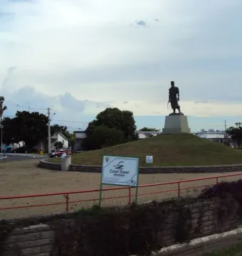 Laçador statue on a pedestal in a traffic circle, with houses, trees, and a cloudy sky behind it.