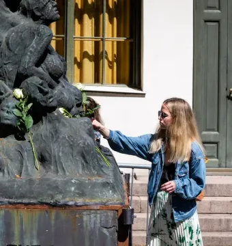Pillar of Shame sculpture with white roses as a woman places another rose on it