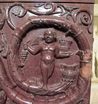 Red porphyry relief on the Sarcophagus of Constantina with a winged putto holding grapes beside a basket, framed by an elaborate vine garland.