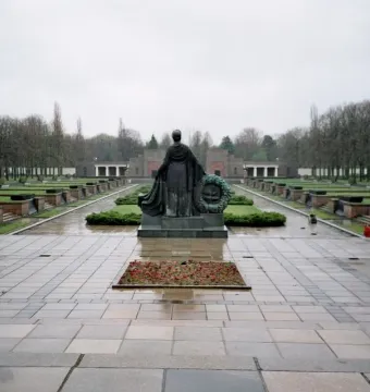 The Soviet War Memorial Schönholzer Heide statue seen from behind, holding a wreath over a wet paved plaza.