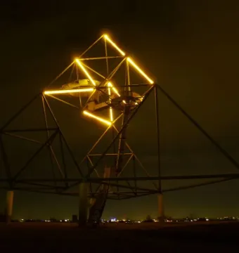 The Tetrahedron sculpture lit at night with yellow lights along its upper edges and a spiral staircase inside.