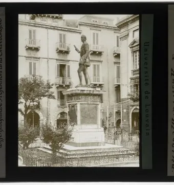 John of Austria monument on a tall ornate pedestal in a public square, with buildings and balconies in the background.