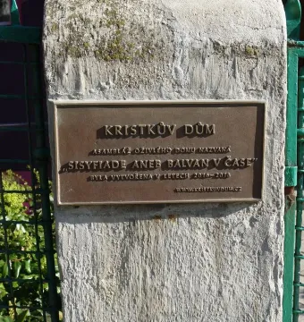Bronze plaque for Kristek House mounted on a light-colored pillar beside a green metal fence.