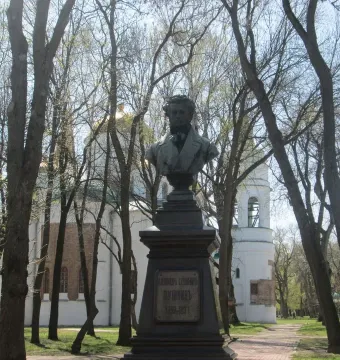 Bust of Pushkin on a dark pedestal in a park with bare trees, a church building, and a bell tower in the background.