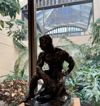The Athlete sculpture by Auguste Rodin displayed on a wooden pedestal with a plaque in front, surrounded by indoor plants and a glass atrium.