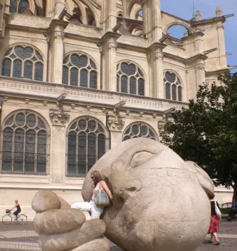 Écoute sculpture showing a large stone head and cupped hand on a plaza, with a woman seated on the hand.