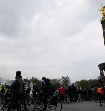 Victory Column in Berlin with a crowd of cyclists gathered around its base under a cloudy sky.