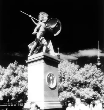 Athena Protects the Young Hero statue on a tall pedestal beside a sidewalk with people walking below.