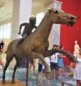 Jockey of Artemision bronze sculpture displayed in a museum with visitors and red walls in the background