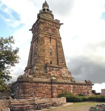 The Kyffhäuser Monument’s Barbarossa Tower rises in reddish-brown stone above a paved forecourt with benches and planting beds.
