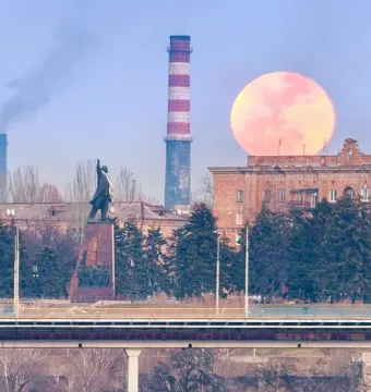 Lenin Monument in Zaporizhzhia with a raised arm, set on a tall pedestal near trees, industrial chimneys, and a large red moon behind a building.