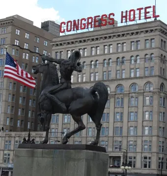 The Bowman and The Spearman bronze equestrian statue on a stone pedestal with a Congress Hotel sign and American flag in the background.