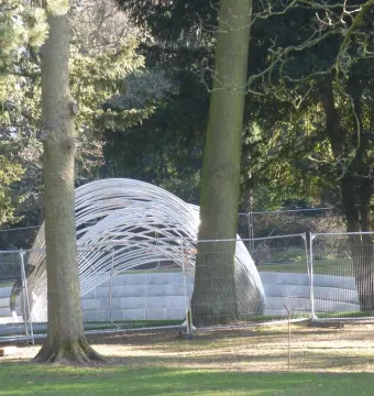 Infinite Wave sculpture in Cannon Hill Park, seen behind a chain-link fence among trees and grass.