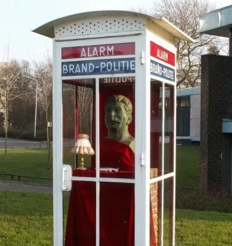 A Stalin bust displayed inside a glass emergency box on a grassy lawn, with a small lamp and fish in front.