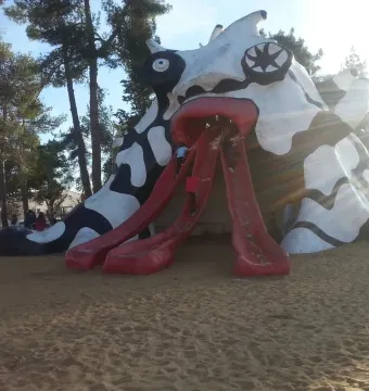 The Golem sculpture with children climbing its three red tongue-like slides in a sandy playground surrounded by trees.