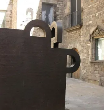 Topos V, a dark geometric sculpture with cutouts, stands in front of historic stone buildings in Barcelona.