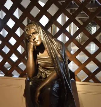 La Douleur bronze sculpture of a seated figure with its head resting on one hand before a wooden lattice screen.