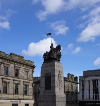 Paisley War Memorial on a tall stone pedestal in a public square, with dark figures on horseback and buildings behind it.