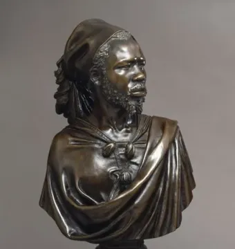 The Nubian Man bronze sculpture shown in three-quarter view on a dark pedestal, with a textured beard, draped garment, and tied head covering.
