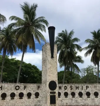 The Torch of Friendship monument’s stone pillar topped with a dark urn and small flame, with palm trees and a low wall behind it.