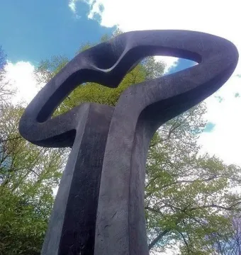 Der Mahner, a dark abstract sculpture with an open loop at the top, framed by green trees against a blue sky.