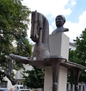 The Statue of Jaroslav Hašek with a bronze-toned horse sculpture on a tall stone pedestal and a bust on a separate pedestal behind it.