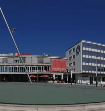 The Man Walking to the Sky sculpture rises on a tilted pole in a plaza in front of the main station buildings.
