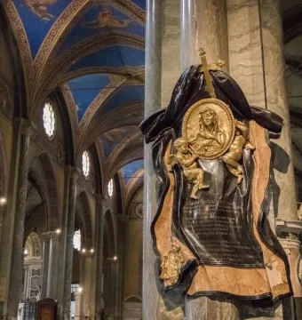 The Memorial to Maria Raggi shows a gilded medallion with a female figure and cherubs mounted on a marble column inside a grand church interior.