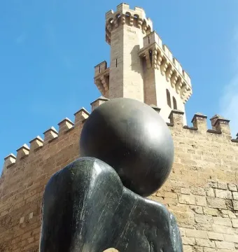 Monument a la dona bronze sculpture with a smooth sphere and undulating base in front of a stone building and tower.