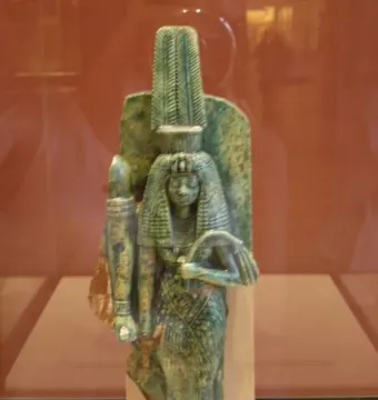 Standing statue of Queen Tiye with a feathered headdress, long wig, and patterned gown, holding a staff-like object.
