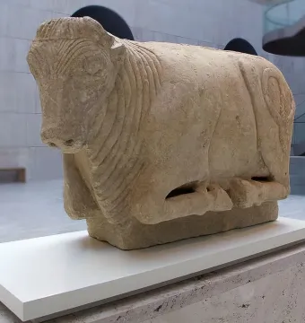 Stone sculpture of the Bull of Osuna in a resting pose on a white display stand.
