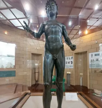 Efebo de Selinunte, a dark bronze nude young man statue with one arm raised in a museum display.