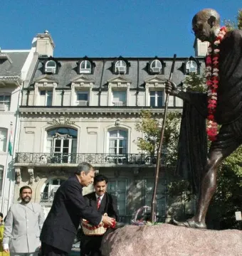 Mahatma Gandhi Memorial statue on a stone boulder with three men standing nearby in front of a building.