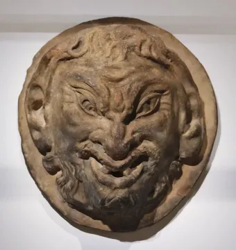 Round relief sculpture of a faun’s head with a wrinkled brow, wide grin, short beard, and pointed ears.