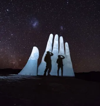 Mano del Desierto sculpture lit against a star-filled night sky, with two people standing in front of it.