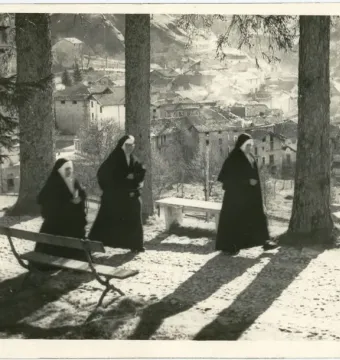 Three nuns walking along a tree-lined path with a village on the hillside in the background.