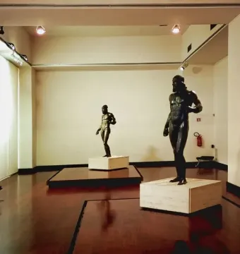 The Riace bronzes displayed on plinths in a museum gallery, with one larger bearded male statue beside a smaller figure.