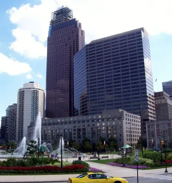 Swann Memorial Fountain spraying high jets in a plaza with skyscrapers and landscaped flower beds nearby.