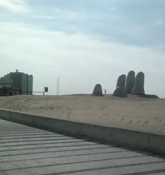 The Hand sculpture with five rounded fingers emerging from a sand dune, with a walkway, cityscape, and ocean in the background.