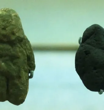 Two rough stone replicas of the Venus of Berekhat Ram, one greenish-brown and figure-like, the other darker and more irregular.