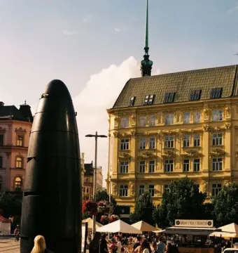 The Brno astronomical clock stands in a sunlit city square with a woman in the foreground and market stalls and buildings behind it.