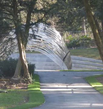 Infinite Wave sculpture spanning a paved walkway in Cannon Hill Park, framed by trees and a fence.