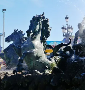 Bronze fountain at the Monument aux Girondins with rearing horses and spraying water, with a fairground attraction and ornate lamppost in the background.